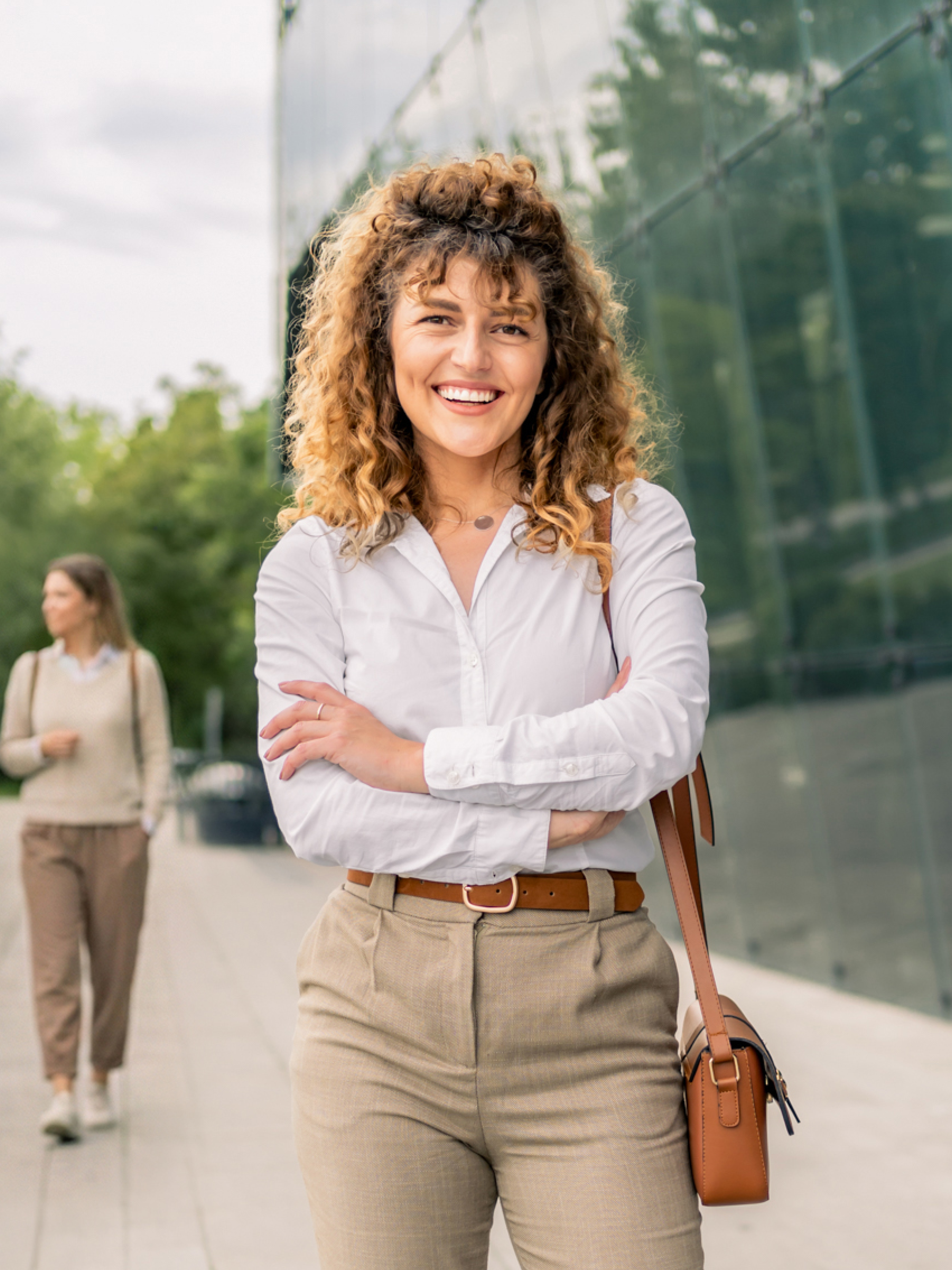 Eine Frau mit Locken im Businesslook verschränkt lachend die Arme. Im Hintergrund ist ein gläsernes Gebäude und Bäume zu sehen.